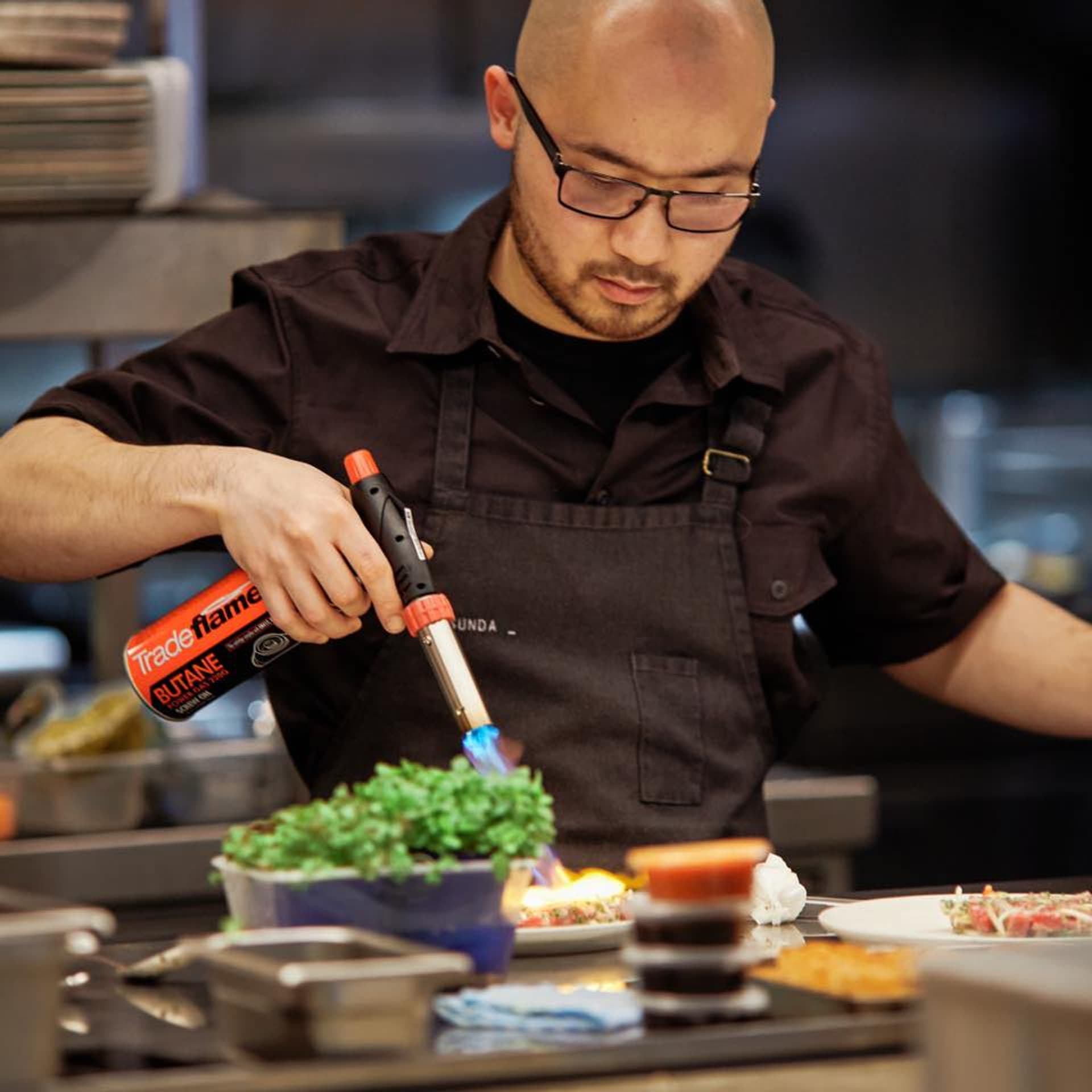 Chef Khanh Nguyen using a kitchen torch to finish a plated dish beside microgreens on a stainless pass
